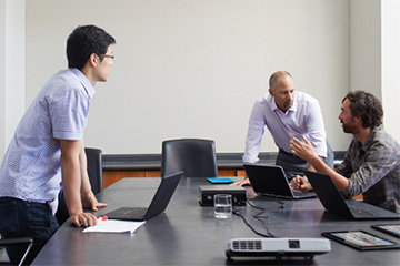 A group of three men sit in a meeting room and talk to each other while using their laptops.